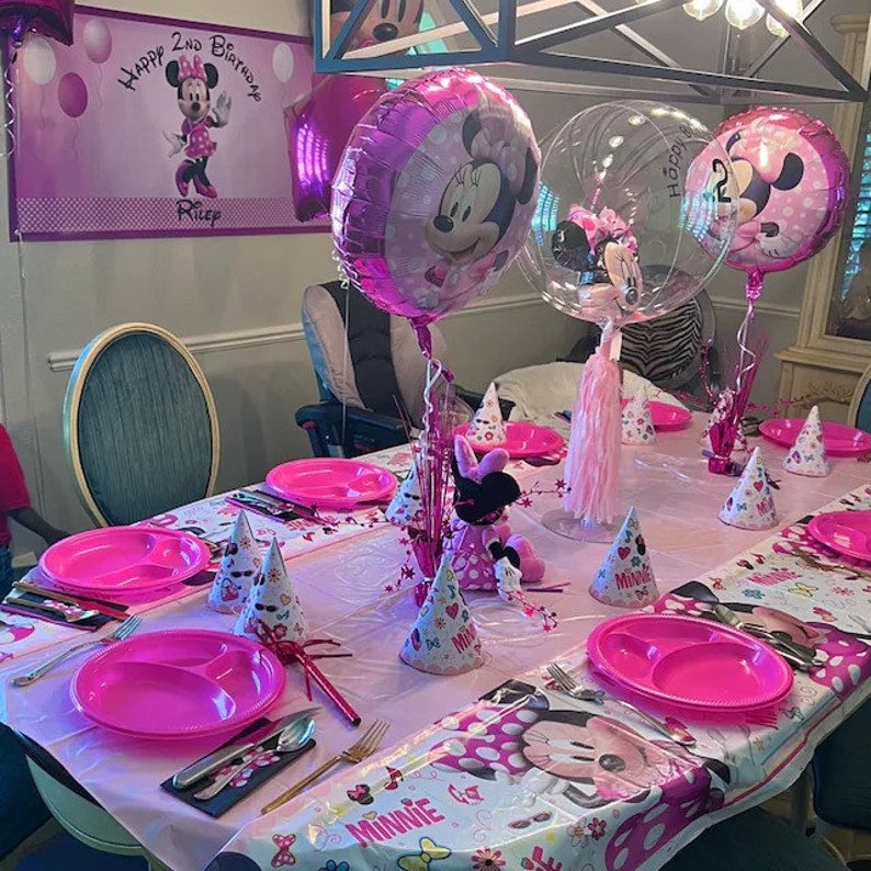 May include: A Minnie Mouse-themed birthday party table setting. Pink plates, party hats, and a Minnie Mouse doll are arranged on a pink tablecloth. Balloons with Minnie Mouse's face and a banner that says "Happy 2nd Birthday" are also visible.