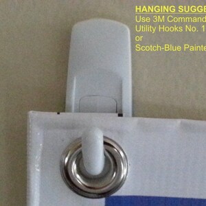 May include: Close-up of a white 3M Command Brand Utility Hook attached to a banner with a metal grommet. The banner has a white border and blue accents. Text on the image suggests hanging options.