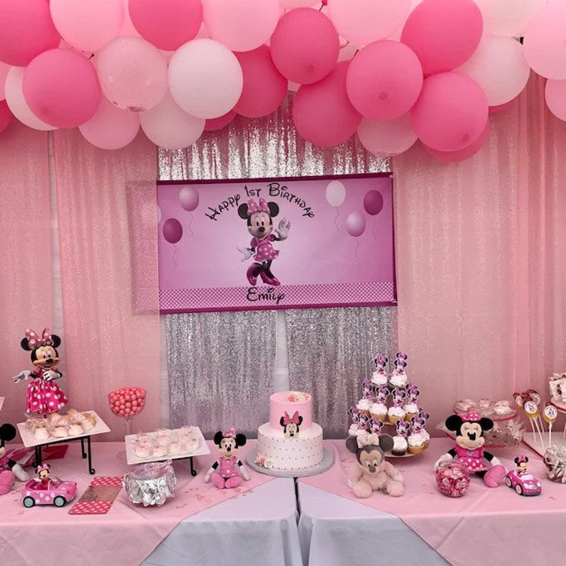 May include: A pink-themed first birthday party setup with Minnie Mouse decorations. The scene includes a banner, cake, cupcakes, balloons, and Minnie Mouse figurines. The banner reads "Happy 1st Birthday" with a name below.