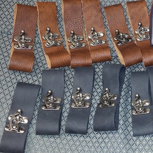 May include: An assortment of leather straps with metal clasps. The straps are in shades of brown and black, with a textured appearance. The metal clasps are silver-toned and add a vintage aesthetic. These straps could be used for various crafting or design projects.