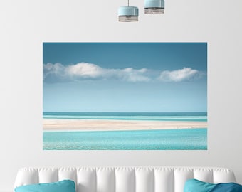 May include: A white couch with blue throw blankets and pillows in a living room with a large print of a beach scene on the wall. The print shows a white sandy beach with turquoise water and a blue sky with white clouds.