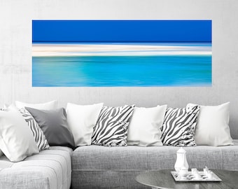 May include: Abstract painting of a blue sky and ocean with white sand. The painting is hanging on a wall above a gray couch with pillows.