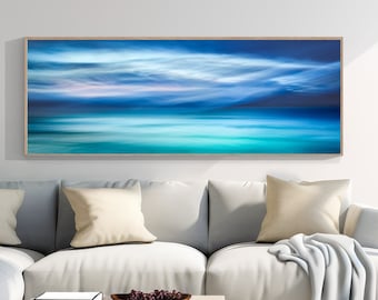 May include: A white sectional sofa with light blue throw pillows and a light blue throw blanket. A framed abstract painting of a blue and green seascape hangs above the sofa. A wooden floor lamp with a white shade stands next to the sofa. A small wooden table with a bowl of fruit sits in front of the sofa.