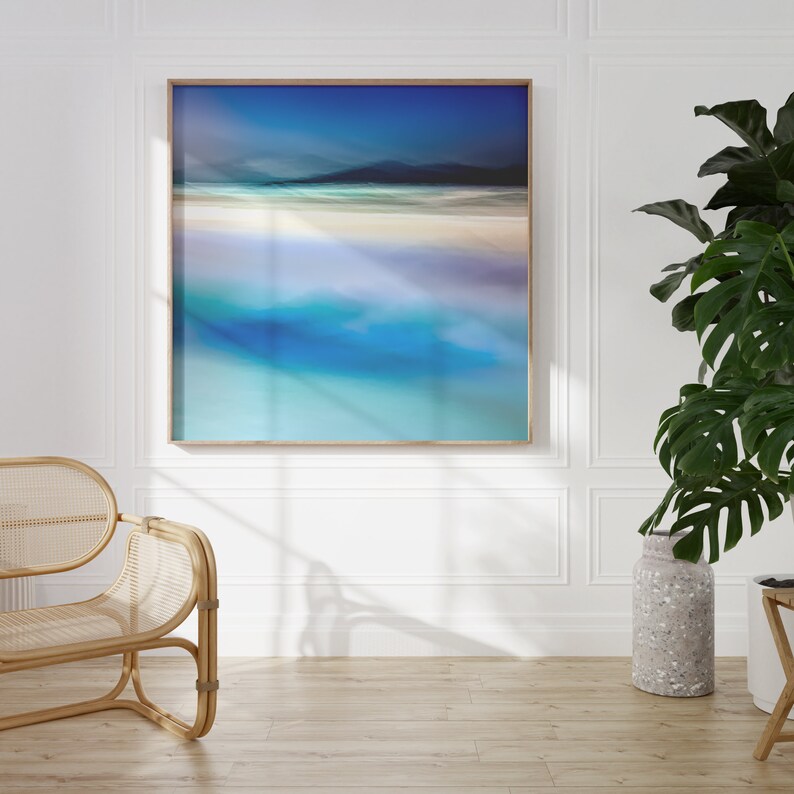 May include: A framed abstract painting of a beach scene with blue and white colours. The painting is in a light wood frame and is hanging on a white wall.