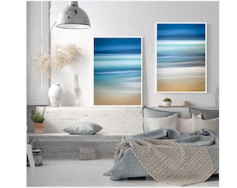 May include: Two framed abstract paintings of a beach scene with blue, white, and brown colors. The paintings are hanging on a white wall above a bed with a gray blanket and pillows.