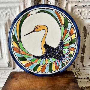 May include: A round ceramic wall plate featuring a hand-painted crane with a yellow neck and head, black body, and multi-colored wings in shades of green, orange, blue, and pink. The plate has a white background and a thick dark blue border.