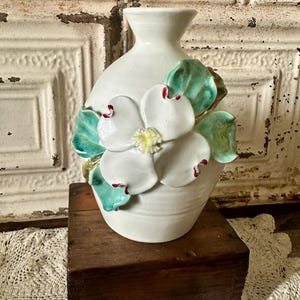 May include: A white ceramic vase with a sculpted dogwood flower design. The flower has white petals with red accents and green leaves. The vase is on a wooden box.