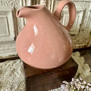 May include: A pink ceramic pitcher with a rounded body and a curved handle. The pitcher has a spout for pouring and sits on a wooden box. The background features a white, textured surface and a sprig of flowers.