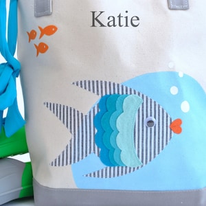May include: A white canvas tote bag with a blue and gray fish design. The bag has a blue ribbon bow and the name "Katie" is printed on the front.