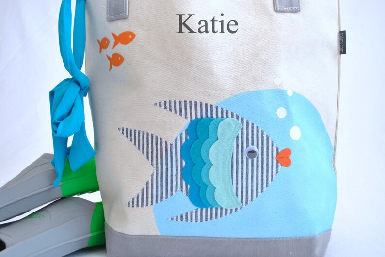 May include: A white canvas tote bag with a blue and gray fish design. The bag has a blue ribbon bow and the name "Katie" is printed on the front.