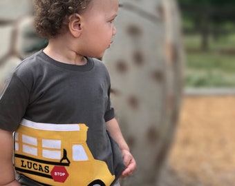May include: A young child wearing a grey t-shirt with a yellow school bus appliqué. The bus has the name "Lucas" and the word "STOP" on it.