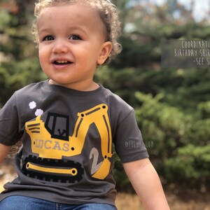 May include: A toddler wearing a gray short-sleeved shirt with a yellow construction vehicle applique. The vehicle has the name "Lucas" and the number "2" on it. The shirt is available in coordinating colors.