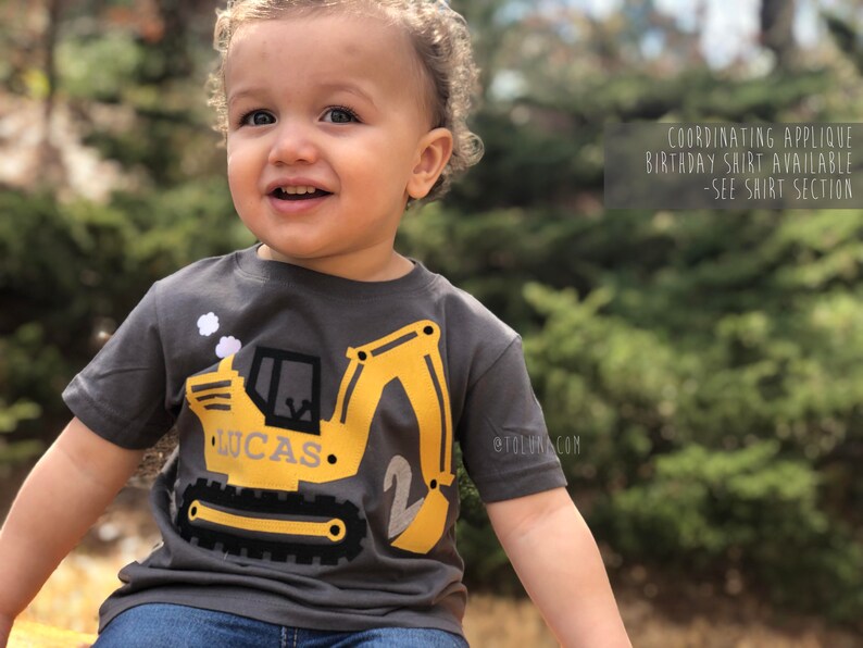 May include: A toddler wearing a gray short-sleeved shirt with a yellow construction vehicle applique. The vehicle has the name "Lucas" and the number "2" on it. The shirt is available in coordinating colors.