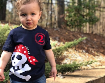 Può includere: Un bambino piccolo che indossa una maglietta blu navy con un design di teschio e ossa incrociate da pirata rosso e bianco. La maglietta ha anche un cerchio rosso con il numero "2" e il nome "Lucas" stampato su di essa.