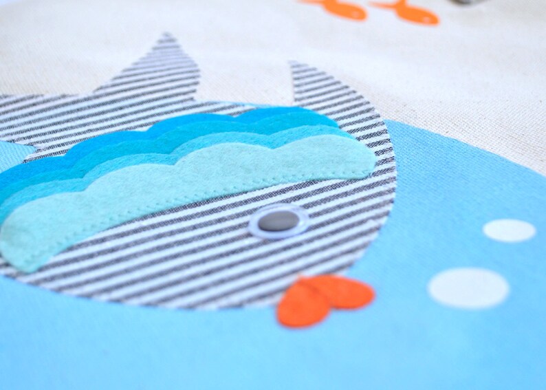May include: A blue and white striped fish with a blue, turquoise, and light blue felt wave design. The fish has a white button eye and a red felt mouth.