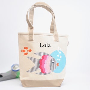 May include: A beige canvas tote bag with a colorful fish design and the name "Lola" printed on the front. The fish has a blue, pink, and gray pattern and is surrounded by orange fish.