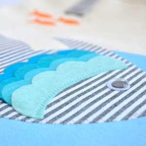 May include: A blue and white striped fabric fish with a blue and green felt fin and a white button eye. The fish is on a blue background.