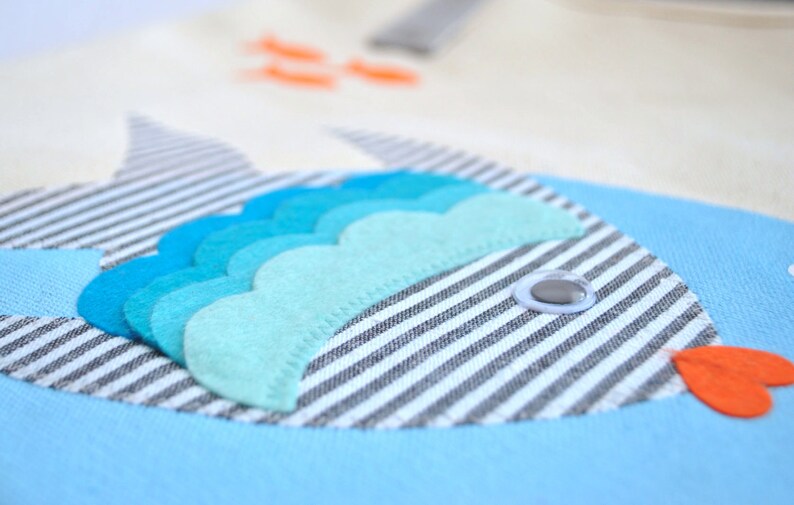 May include: A blue and white striped fabric fish with a blue and green felt fin and a white button eye. The fish is on a blue background.