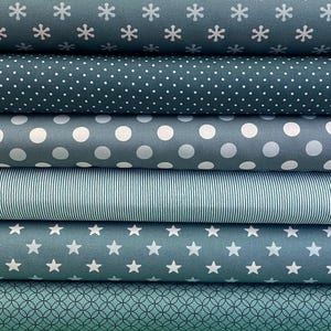 Könnte beinhalten: Ein Stapel verschiedener Stoffe in Teal- und Grautönen. Die Muster umfassen Schneeflocken, Punkte, große Kreise, vertikale Streifen, Sterne und ein geometrisches Design. Die Stoffe sind ordentlich gefaltet und zeigen verschiedene Texturen und Designs.