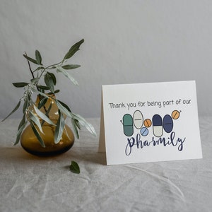 May include: A greeting card with the text "Thank you for being part of our pharmily." The card features a design of cartoon pills in various colors. A brown glass vase with green leaves is in the background.