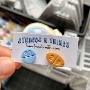 May include: A pair of pill-shaped earrings, one blue with the number 30 on it, and one orange with the number 20 on it. The earrings are on a white card with the text "STRINGS N THINGS handmade with love".