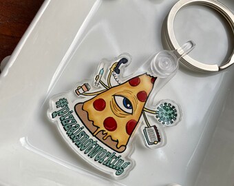 May include: A clear acrylic keychain featuring a cartoon pizza slice with a single eye, holding a phone, coffee cup, syringe, and a laptop. The pizza slice is dripping with cheese and has the text "#pizzainotworking" on the bottom.