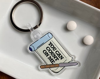 May include: A white and blue keychain with a rolled up scroll that says "You can count on me" and a small knife. The keychain is on a white ceramic dish with two white pills.