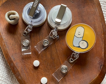 May include: Three clear plastic badge holders with retractable reels. The holders have a silver clip and a round, clear plastic insert. One holder has a yellow background with a cartoon illustration of a pill with a smiling face and a smaller pill with a smiling face.