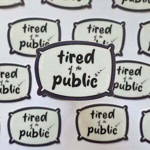 May include: A set of nine white pillow-shaped stickers with black outlines. Each sticker features the text "tired of the public" with a stylized "zzz" at the end. The stickers are arranged in a grid pattern on a white surface.