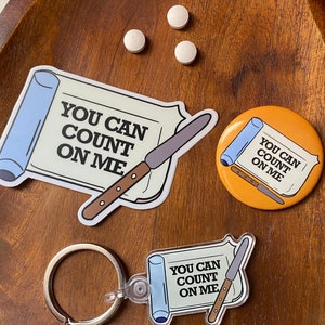 May include: Three different versions of a sticker with the text "You can count on me" and a knife on a scroll. The sticker is available as a sticker, a button, and a keychain.