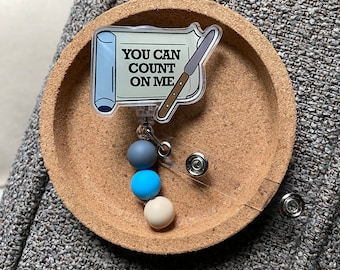 May include: A white and blue badge reel with a paper scroll design that says "You can count on me". The scroll has a knife design on it. The badge reel has three beads: one gray, one blue, and one cream.