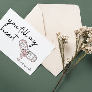 Puede incluir: Una tarjeta blanca con el texto "you fill my heart - and my scripts" y un dibujo de una cápsula de píldora llena de corazones. La tarjeta está sobre una superficie verde con un sobre blanco y una rama de flores blancas.