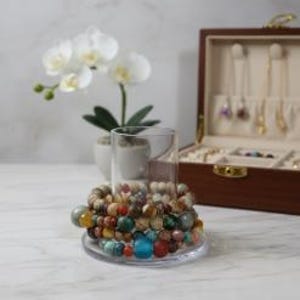 May include: A stack of multi-colored gemstone bead bracelets featuring various earth tones, including shades of blue, red, brown, and green, wrapped around a clear glass cylinder. The beads vary in size and texture, showcasing natural stone patterns.