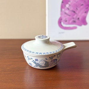 May include: A white ceramic lidded serving dish with blue floral patterns and a handle. The dish has a round shape with a lid and a small handle on the side. The floral design includes blue flowers and leaves.