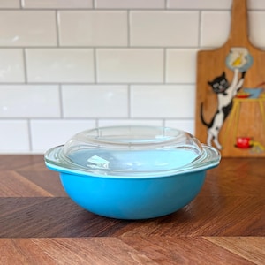 May include: A bright turquoise round glass bowl with a clear glass lid. The bowl features a smooth, solid-colored exterior and a wide rim. The lid is transparent, domed, and fits securely over the top of the bowl, creating a complete kitchen storage container.