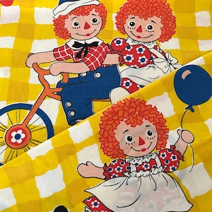 Op de afbeelding: Stof met een geel en wit geruit patroon met de poppen Raggedy Ann en Andy. Eén pop fietst, terwijl een andere een blauwe ballon vasthoudt. De poppen hebben rood garen haar en zijn gekleed in vintage kleding.