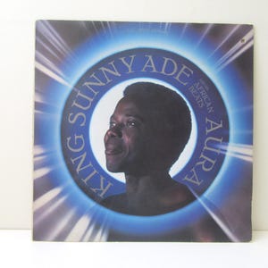May include: Album cover for King Sunny Ade's "Aura" featuring a portrait of the musician. The album art has a blue and white circular design with the artist's name and album title. The words "African Beats" are also visible.