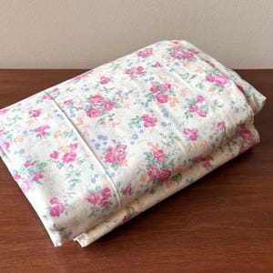 May include: Folded floral bed linen. The sheets have a cream background with a pattern of pink roses, blue accents, and green leaves. The fabric is neatly folded on a wooden surface.