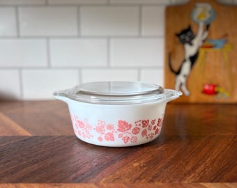 Puede incluir: Una fuente de horno de vidrio blanco con tapa de vidrio transparente, que presenta un patrón repetitivo de hojas y frutos de grosella espinosa en color rosa alrededor del exterior. El recipiente redondo tiene dos asas integradas.