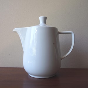May include: A white ceramic jug with a lid. The jug features a curved handle and a spout for pouring. The lid is topped with a small knob. The jug is placed on a dark brown wooden surface.