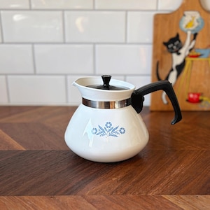 May include: White vintage teapot with a black handle and lid, and a silver band. The teapot has a floral design in blue. The teapot is on a wooden surface with a white tile background.
