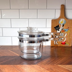 May include: A clear glass double boiler with a lid and silver handles. The kitchenware sits on a wooden surface with a decorative cutting board in the background. The cutting board has a cat illustration.