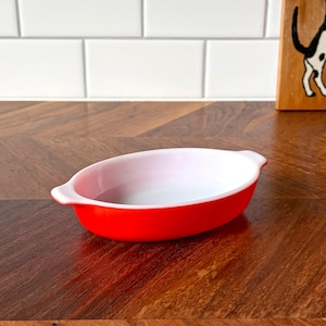May include: An oval red and white baking dish with handles. The dish has a bright red exterior and a white interior. The dish is on a wooden surface.