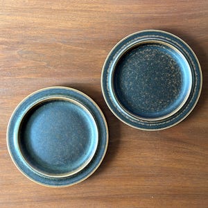 May include: Two round ceramic plates with a dark teal centre and a lighter tan rim. The plates have a speckled texture and are set against a wooden surface. The plates are likely for serving food.