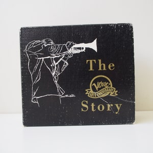 Könnte beinhalten: Schwarze Box mit einer weißen Illustration einer Person, die Trompete spielt. Die Box trägt einen goldenen Schriftzug "The Verve 50th Anniversary Story".