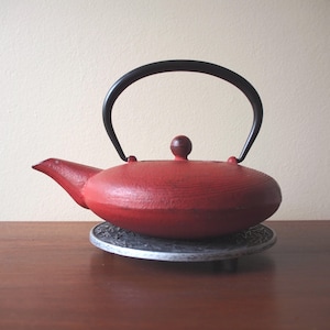 May include: A red cast iron teapot with a black handle and a small, round lid. The teapot sits on a decorative metal trivet. The teapot has a spout for pouring and is designed for brewing tea.
