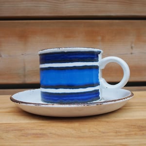 Könnte beinhalten: Eine Keramik-Tasse mit passender Untertasse. Die Tasse hat horizontale Streifen in Blau, Weiß und Schwarz. Der Rand und der Henkel sind weiß. Die Untertasse ist weiß mit einem braunen Rand. Das Set steht auf einer Holzoberfläche.