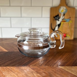 May include: A clear glass teapot with a silver band around the middle and a curved handle. The teapot has a lid and a spout. The background includes a wooden cutting board with a cat illustration and white tiles.