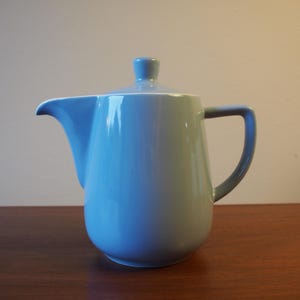 Peut inclure: Une théière en céramique bleu clair avec un couvercle et une poignée assortis. La théière a un bec courbé et un corps arrondi. La poignée est d'une nuance de bleu plus foncée. La théière est sur une surface en bois.