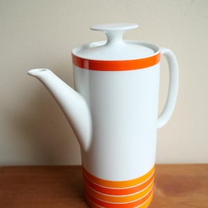 May include: A white ceramic coffee pot featuring a cylindrical body, a curved handle, and a spout. The design includes a solid orange band near the top rim and four horizontal orange stripes encircling the base, topped with a white lid.
